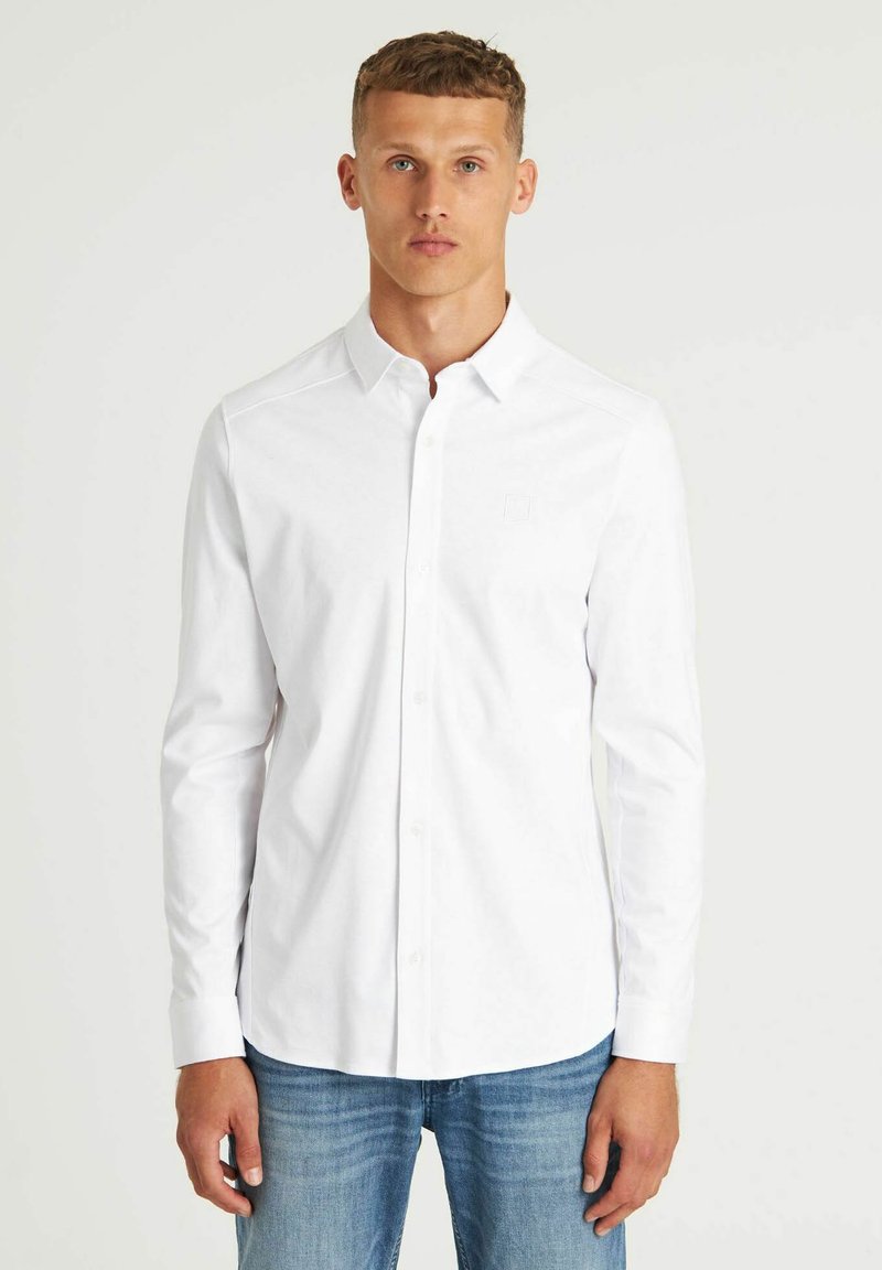 Weißes langärmliges Hemd mit Button-Down-Kragen, aus Baumwolle, mit einem dezenten Logo auf der Brust und geradem Saum. Getragen mit blauen Jeans.