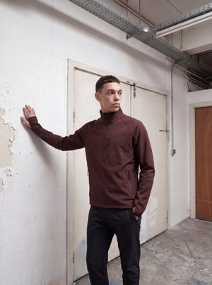 Jeune homme en veste marron zippée, la main posée sur un mur blanc qui s'écaille, debout dans une pièce industrielle usée avec un sol en béton et des luminaires métalliques au plafond.