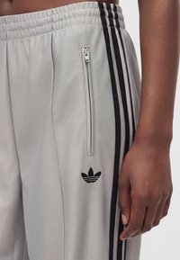 Κοντινή λήψη γκρι αθλητικών παντελονιών Adidas με μαύρες τρίπλες ρίγες, τσέπη με φερμουάρ και κεντημένο λογότυπο στο ισχίο και το χέρι ενός ατόμου.