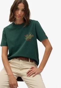 Grøn bomulds t-shirt med korte ærmer, der har et guld- og rødt grafisk design. Parret med beige bukser og et flettet brunt bælte.