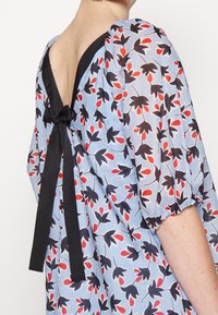 Jasnoniebieska sukienka w kwiaty z bufkami na rękawach, otwartymi plecami związanymi czarną kokardą, odsłaniającymi górną część pleców i ramiona.