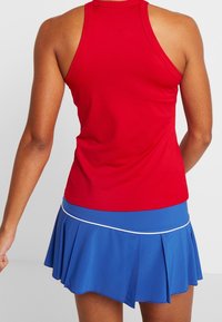 Débardeur de sport rouge avec un dos nageur, associé à une jupe plissée bleue avec des bordures blanches.