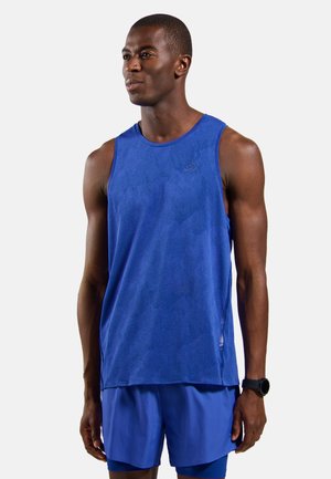 Atletische man in een blauw mouwloos tanktop en korte broek staand, met een zwart polshorloge, kijkt iets naar links op een lichtgrijze achtergrond.