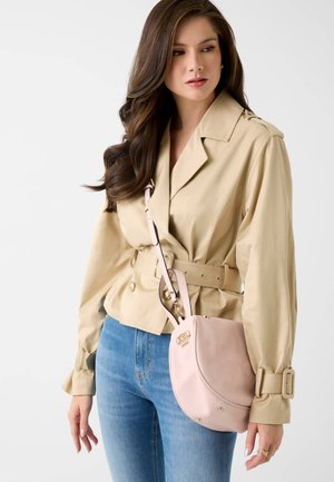 Woman wearing beige belted jacket and blue jeans, carrying a light pink shoulder bag with gold accents.