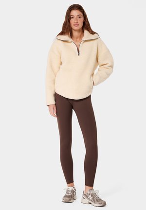 Młoda kobieta stoi, ubrana w kremowy polar z półzamkiem, ciemnobrązowe legginsy i beżowe buty do biegania na jednolitym tle.