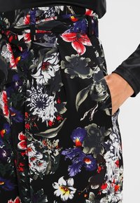 Floral print trousers featuring a black background, intricate multicolored flowers, a tie waist, and side pockets. Soft fabric texture.