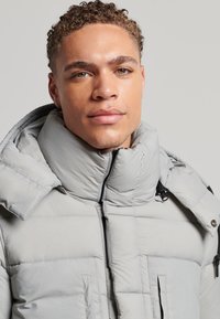 Chaqueta acolchada gris claro con cuello alto, cierre de cremallera y dos bolsillos en el pecho. Presenta una textura suave y un patrón de cuadrícula en la tela.