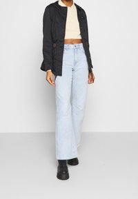 Chaqueta negra corta con capucha y cordón, camiseta corta beige claro, jeans acampanados azul claro y botas de plataforma negras con cordones.