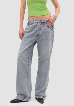 Szczupła talia w jasnych, szerokich dżinsach, limonkowy crop top, srebrne pierścionki, bransoletka i czarne, spiczaste buty na jednolitym tle.