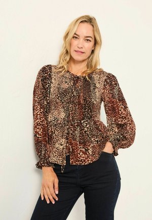 Blonde Frau in einer langärmligen, gemusterten Bluse in Braun- und Schwarztönen, mit dunklen Hosen, vor einer schlichten hellen Wand stehend.