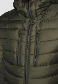 Veste matelassée verte avec fermeture éclair à l'avant, deux poches poitrine avec zippers et une capuche. Elle présente un tissu texturé et des accents en métal noir.