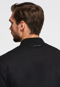 Czarna kurtka wykonana z teksturowanej tkaniny, z wysokim kołnierzem i charakterystycznym detalem zapięcia z tyłu. Materiał wydaje się być strukturalny i trwały.