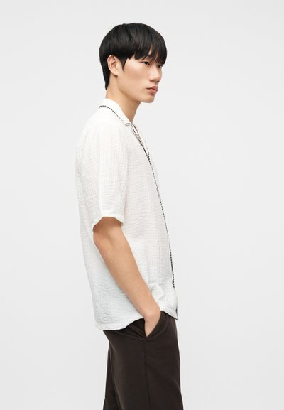 Νεαρός άνδρας με λευκό πουκάμισο κοντομάνικο με υφή και μαύρη λεπτομέρεια, με τα χέρια στις τσέπες, στέκεται μπροστά σε απλό φωτεινό φόντο, κοιτάζοντας προς τα αριστερά.
