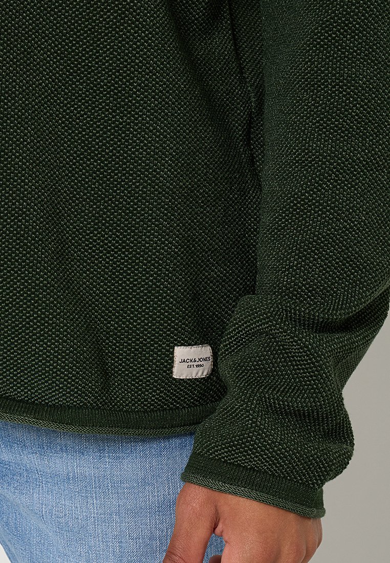 Donkergroen gebreide trui met een gestructureerde weving. Heeft ribboorden bij de mouwen en onderkant, met een klein merkembleem aan de onderzijde.