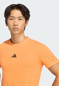 Camiseta deportiva naranja hecha de tela suave, con cuello redondo, mangas cortas y un logo de Adidas negro en el pecho.