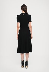 Black ribbed dress with short sleeves, fitted bodice, flared skirt, and a subtle gold accent at the back. Paired with black heeled ankle boots.