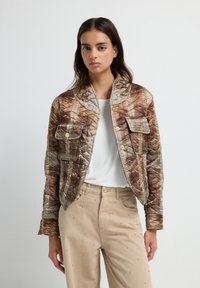 Quiltet jakke i jordfarver med et mønstret design, der har en krave, to frontlommer og knapper i guld-tone. Parret med beige bukser.