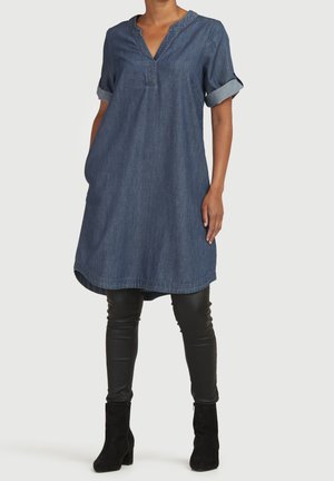 Kvinde iført en blå denim knælængde kjole med op-rullede korte ærmer over sorte tætsiddende læderbukser og sorte ankelstøvler.