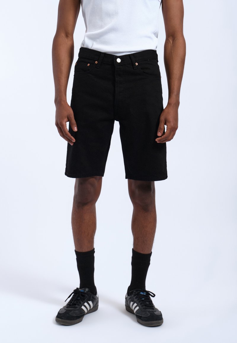 Pantalones cortos de mezclilla negra con un ajuste regular, que cuentan con un cierre de botón y cremallera, bolsillos delanteros y una textura suave, que se llevan con calcetas deportivas negras y zapatillas.