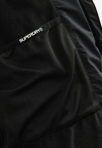 Juoda striukė, pagaminta iš minkštos, tekstūruotos vilnos ir lygaus, blizgaus audinio, su tinkleliu ir "SUPERDRY" logotipo detale.