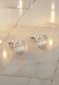 Silverfärgade cirkulära örhängen med intrikata utklippta geometriska mönster. Matt yta med polerad kant, fäst med en stud och fjärilsklaff.