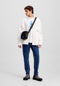 Sacoche noire en bandoulière avec un petit accent bleu, portée sur une chemise blanche et un jean en denim clair, complétée par des chaussures gris à enfiler.
