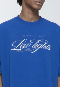 Modrá bavlněná trika s velkým bílým grafickým potiskem s kurzívním textem. Design obsahuje "Low Lights" a další textové detaily.