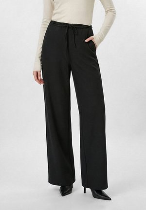 Pantalones negros de pierna ancha con cintura de cordón, combinados con una blusa beige de manga larga y zapatos negros de tacón alto con punta puntiaguda.