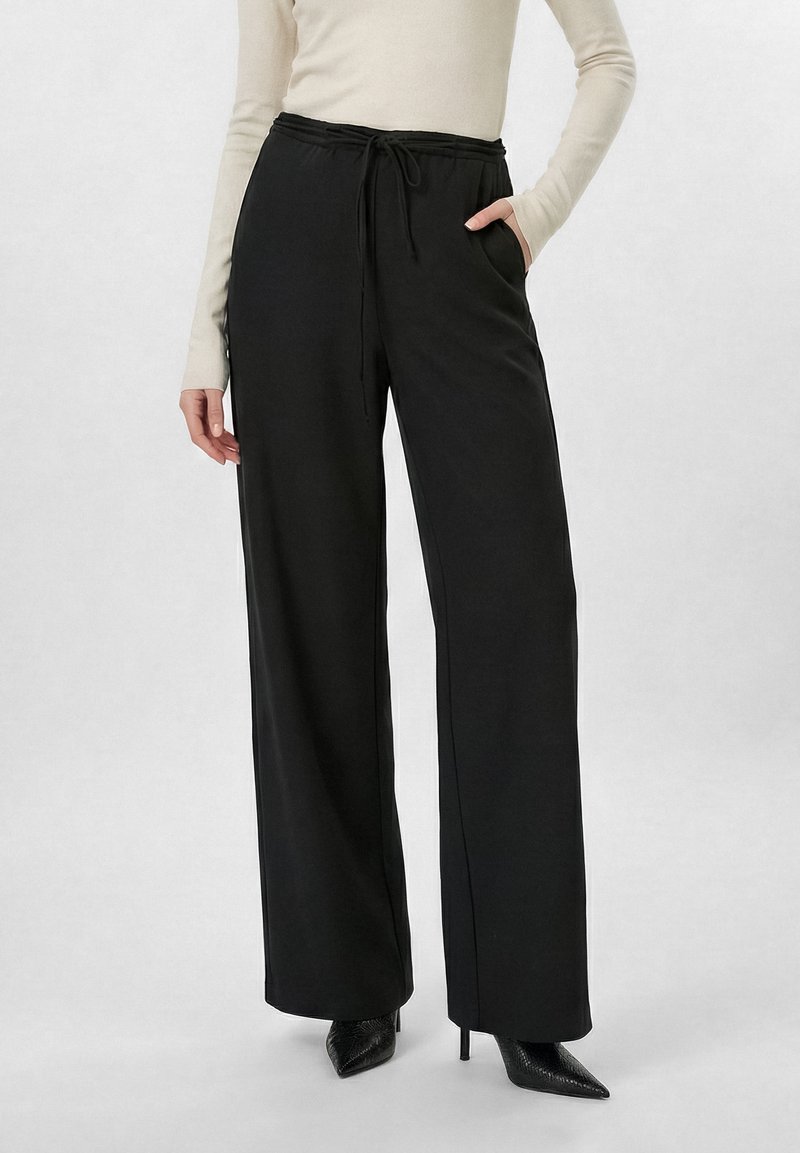 Μαύρο παντελόνι φαρδιάς γραμμής με σούρα στη μέση, φορεμένο με μπεζ μπλούζα μακρυμάνικη και μαύρα μυτερά ψηλοτάκουνα παπούτσια.