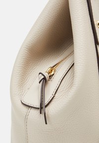 Close-up van een beige leren tas met een gouden rits en een leren trekband die in een knoop is gestrikt.