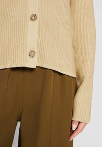 Cardigan en tricot beige avec boutons écaille de tortue, assorti à un pantalon large marron taille haute, montrant la main droite et le poignet.