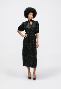 Žena s afro frizurom nosi crnu midi haljinu s šljokicama i balon rukavima, u crnim visokim potpeticama, stoji s jednom rukom na kuku.