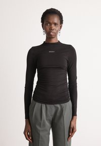 Black long-sleeve top featuring a ruched design and a subtle logo at the chest, paired with loose gray trousers for a contrast in style.