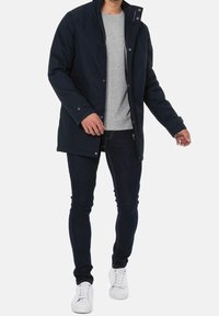 Mężczyzna w granatowej kurtce noszący szary T-shirt, ciemne skinny jeans i białe trampki, stojący na białym tle.