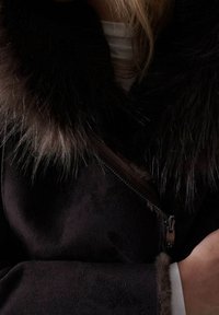 Abrigo marrón oscuro con grueso cuello y puños de piel, usando una blusa de color claro por debajo, con el brazo y la mano visibles descansando cerca del cuerpo.