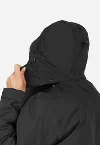 Svart jacka med en välsittande huva, justerbar dragsko och knappknäppning; har en slät yta och minimalistisk design.