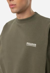 Khaki sweatshirt with a crew neck and ribbed trim. Features a white "PEGADOR" logo on the left chest, showcasing a smooth texture.