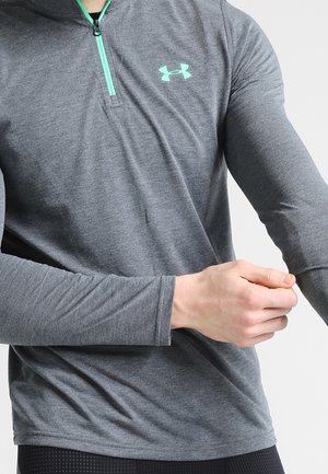 Mann passt Ärmel eines grauen Langarm-Sportshirts mit türkisfarbenem Reißverschluss und Logo auf der Brust an.