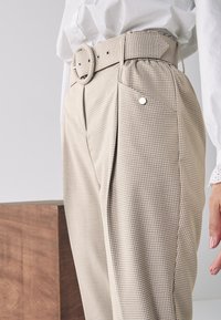 Beige geruite broek met een verstelbare taille en zijzakken. Heeft een gestructureerde stof en een knoopaccent nabij de taille.