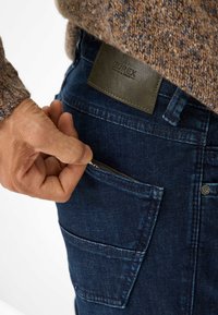 Jeans in denim blu scuro con una tasca posteriore, caratterizzati da una toppa in pelle verde con testo impresso. Maglione strutturato parzialmente visibile sopra.