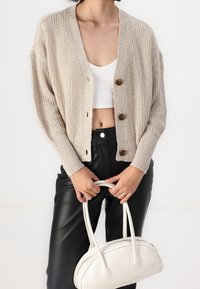 Beige stickad cardigan med V-ringning, två stora knappar och ribbstickade manschetter, i kombination med högmidjade svarta fusk läderbyxor och en vit handväska.