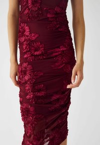 Burgundy figurnära klänning med blommig broderi. Har rynkad textur, intrikata blommönster och ett mjukt, stretchigt tyg.