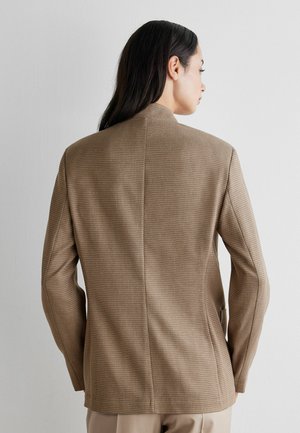Kvinde med langt mørkt hår iført en tekstureret brun blazer, set bagfra mod en ensartet lys væg.