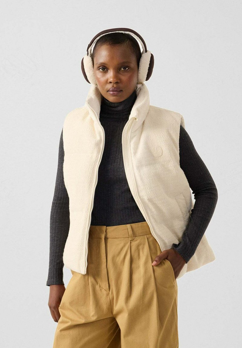 Cremefarbene Fleeceweste mit hohem Kragen, Reißverschluss und strukturiertem Muster, kombiniert mit einem schwarzen Rollkragenpullover und beigefarbenen Hosen. Braune Ohrenschützer getragen.