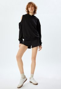 Sudadera negra con corte en el hombro, combinada con pantalones cortos negros. El conjunto está hecho de un material suave y tiene un ajuste relajado. Zapatillas de color claro con acentos en beige.