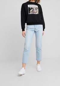 Felpa nera con stampa grafica raffigurante il volto di una donna e del testo. Abbinata a jeans a vita alta di colore blu chiaro e sneakers bianche.