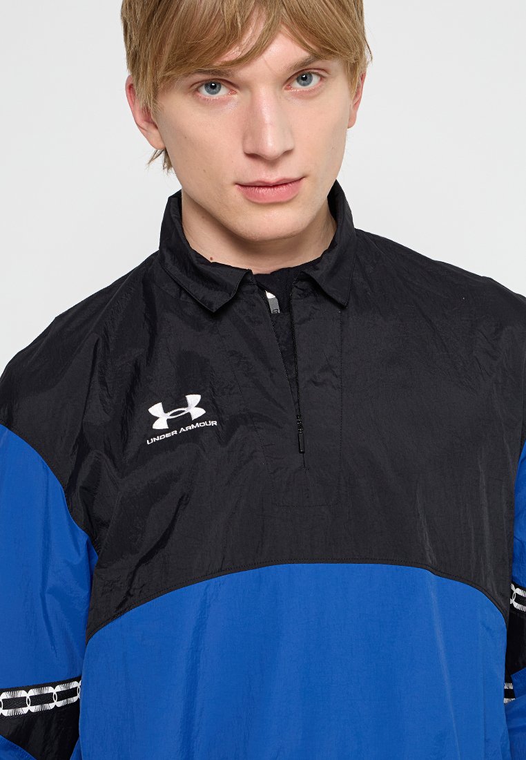 Chaqueta de abrigo negra y azul con cuello con cremallera, fabricada con un material suave. Presenta el logo de Under Armour y detalles a rayas en las mangas.