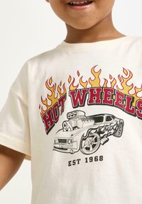 Kermanvärinen puuvillapaita, jossa on punaisella ja mustalla "Hot Wheels" -teksti, sekä grafiikka autosta ja liekkikoristeita oranssina ja keltaisena.