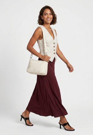 Donna sorridente cammina indossando un gilet senza maniche abbottonato, gonna bordeaux, sandali neri con tacco e porta una borsa a tracolla bianca.