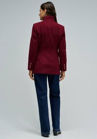 Blazer entallado burdeos con un cuello alto, mangas largas y acentos de botones. Lleva puesto sobre jeans ajustados oscuros, mostrando un corte estructurado.